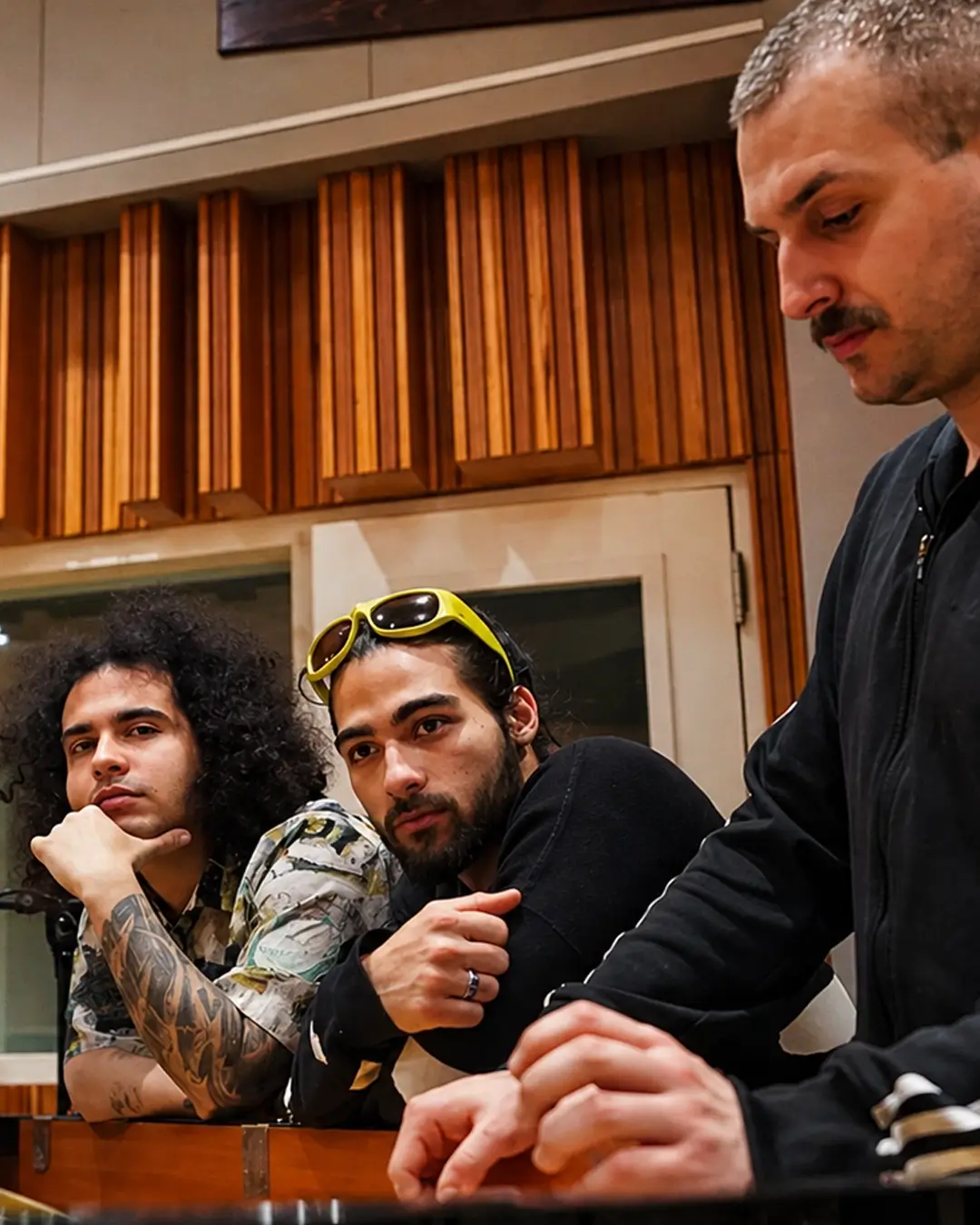 Vier muzikanten staan rond een piano in een opnamestudio, geconcentreerd en in gesprek, met warme houten panelen op de achtergrond.