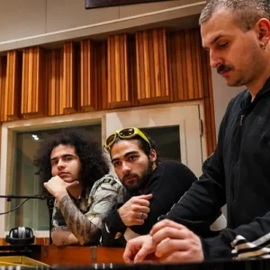 Vier muzikanten staan rond een piano in een opnamestudio, geconcentreerd en in gesprek, met warme houten panelen op de achtergrond.