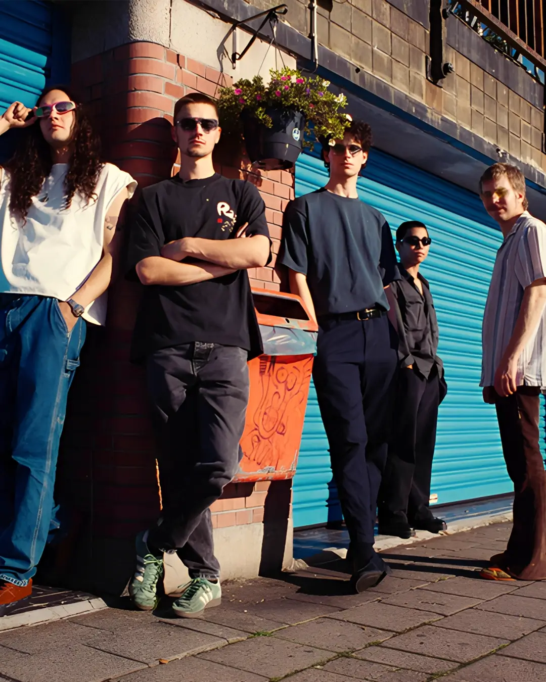 Vijf jonge muzikanten staan ontspannen tegen een muur met blauwe rolluiken, in zonlicht, als bandportret in een stedelijke omgeving.