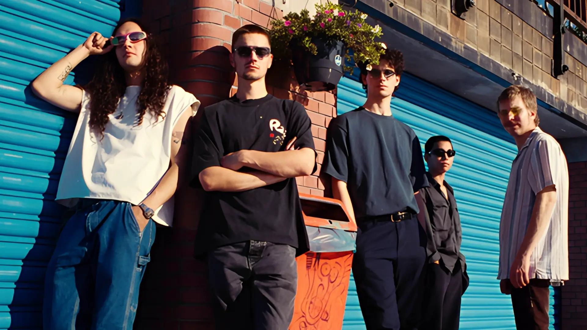 Vijf jonge muzikanten staan ontspannen tegen een muur met blauwe rolluiken, in zonlicht, als bandportret in een stedelijke omgeving.