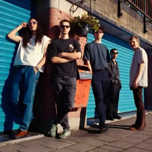 Vijf jonge muzikanten staan ontspannen tegen een muur met blauwe rolluiken, in zonlicht, als bandportret in een stedelijke omgeving.