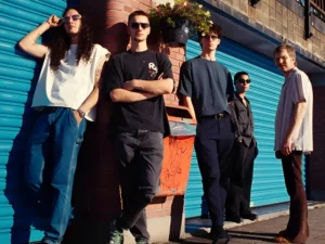 Vijf jonge muzikanten staan ontspannen tegen een muur met blauwe rolluiken, in zonlicht, als bandportret in een stedelijke omgeving.