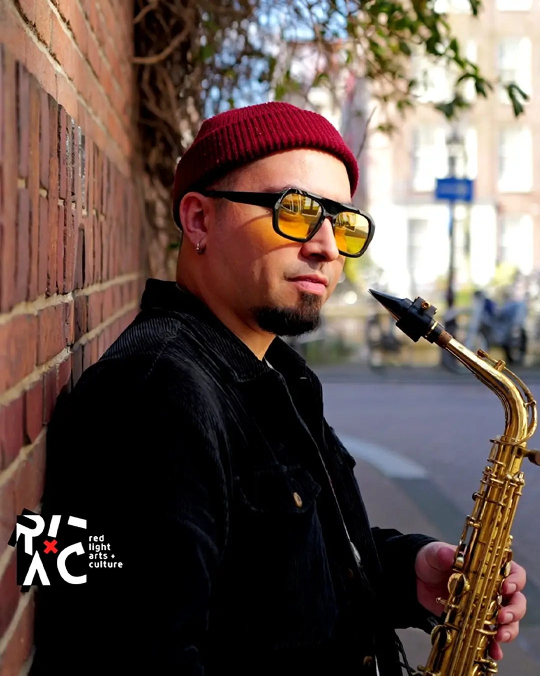 Saxofonist César Vidal van het César Vidal Trio leunt tegen een bakstenen muur en houdt zijn saxofoon vast, gekleed in een rode muts en zonnebril op een zonnige straat.