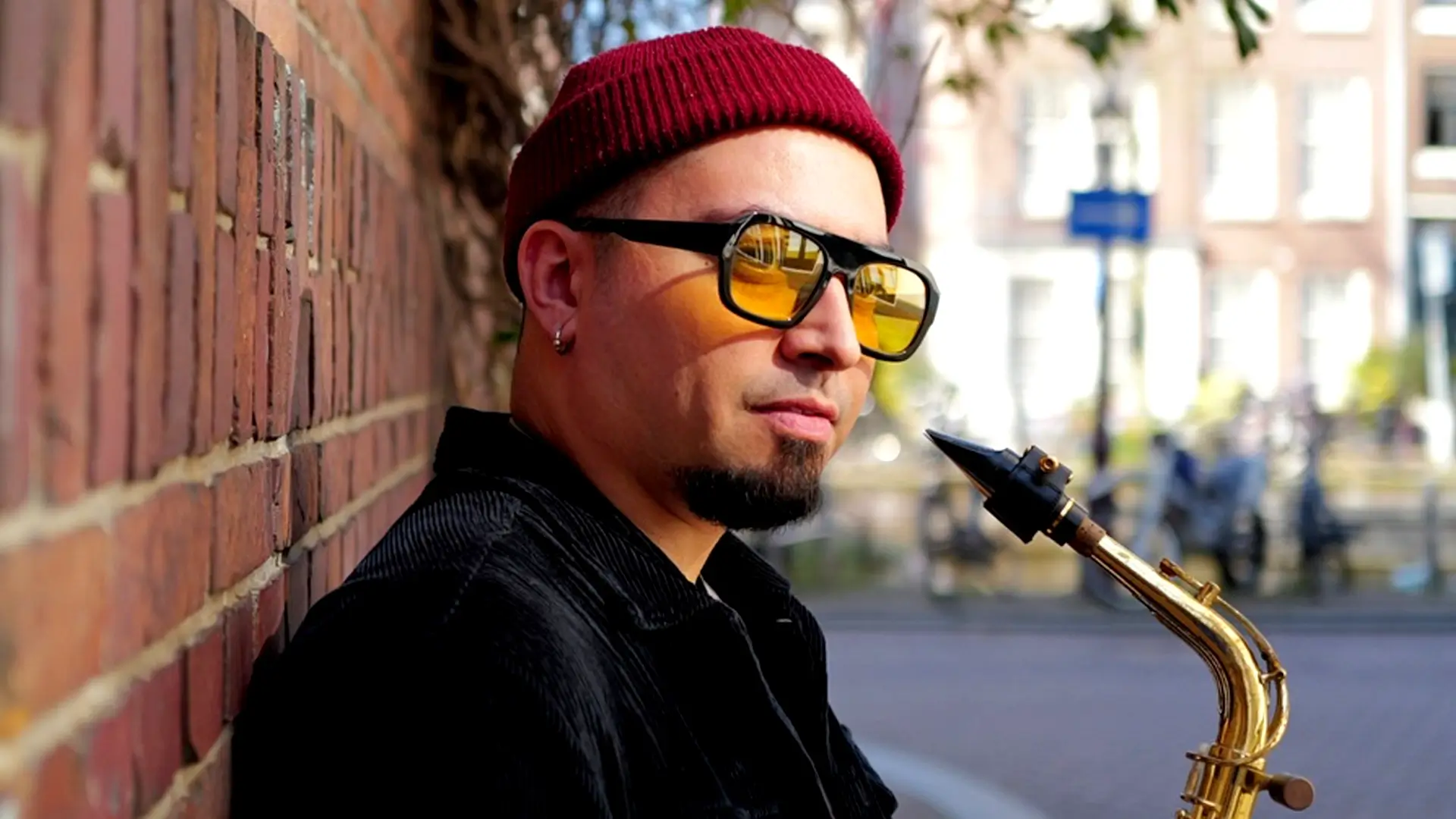 Saxofonist César Vidal van het César Vidal Trio leunt tegen een bakstenen muur en houdt zijn saxofoon vast, gekleed in een rode muts en zonnebril op een zonnige straat.