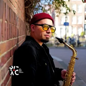 Saxofonist César Vidal van het César Vidal Trio leunt tegen een bakstenen muur en houdt zijn saxofoon vast, gekleed in een rode muts en zonnebril op een zonnige straat.