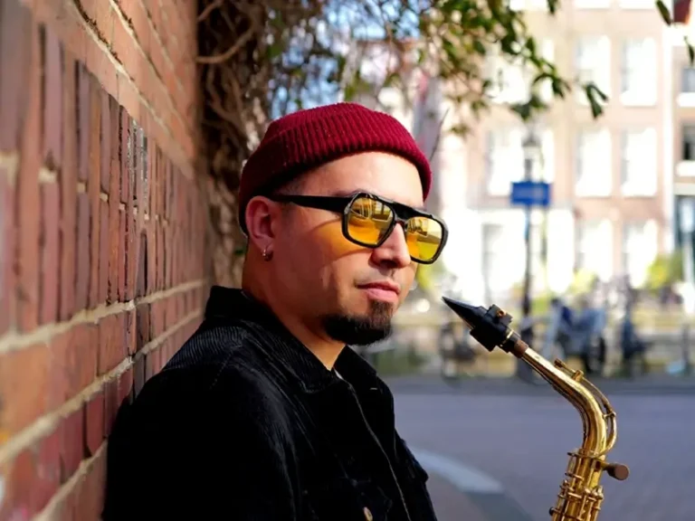 Saxofonist César Vidal van het César Vidal Trio leunt tegen een bakstenen muur en houdt zijn saxofoon vast, gekleed in een rode muts en zonnebril op een zonnige straat.