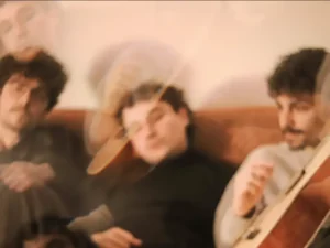 Three musicians sit together on a couch in a warm, dreamy, slightly blurred setting.