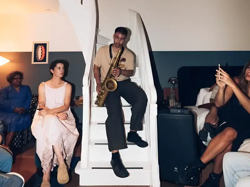 Saxofonist Mark Beebe zit op een trap en speelt terwijl mensen ontspannen in een huiselijke setting om hem heen zitten, sommigen luisterend en anderen met een drankje of telefoon in de hand.
