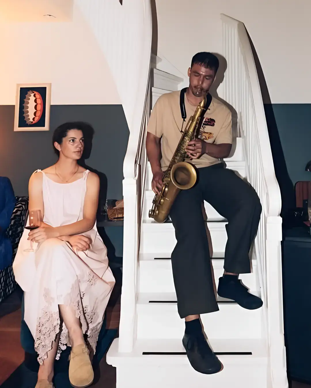 Saxofonist Mark Beebe zit op een trap en speelt terwijl mensen ontspannen in een huiselijke setting om hem heen zitten, sommigen luisterend en anderen met een drankje of telefoon in de hand.