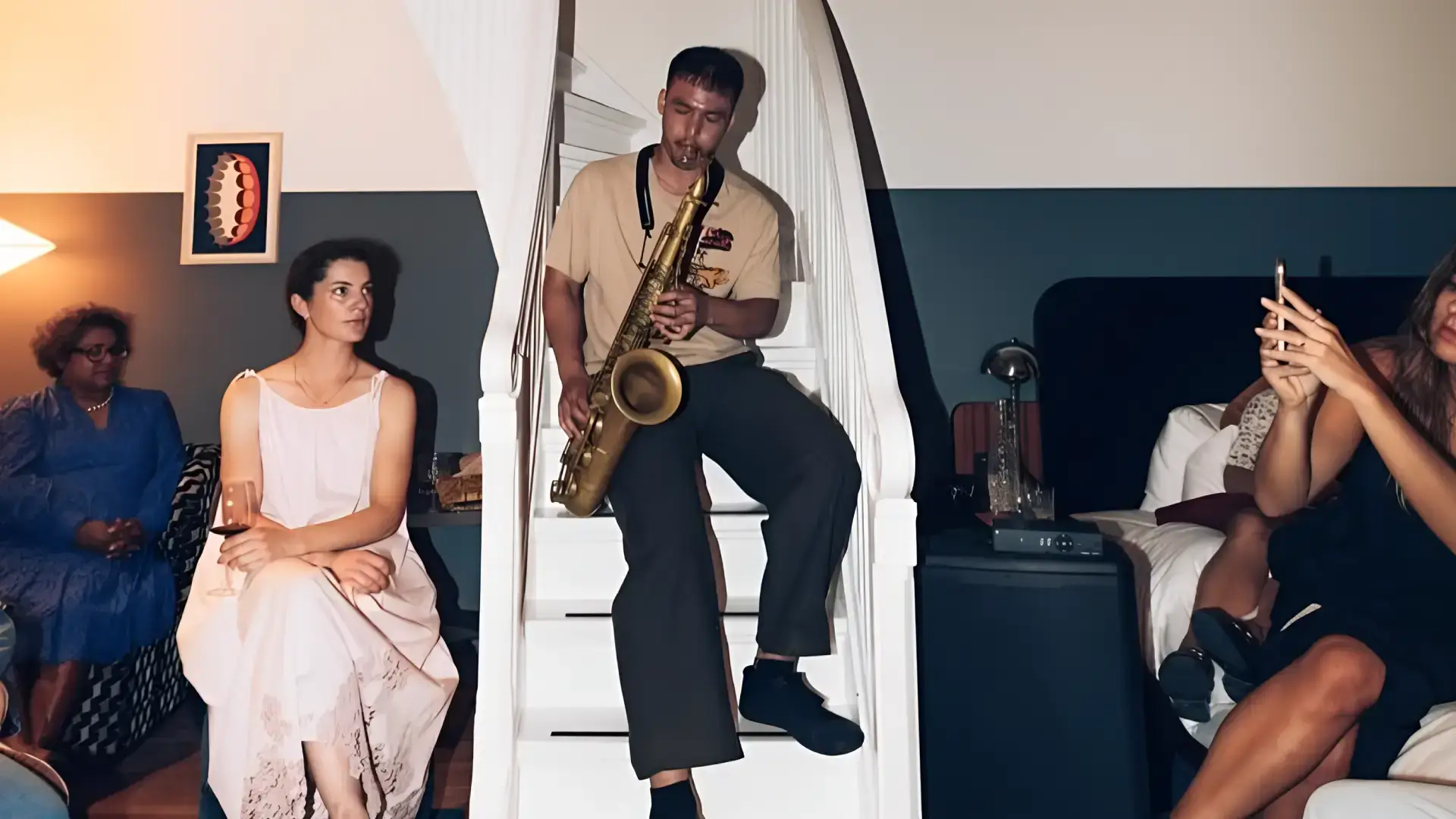 Saxofonist Mark Beebe zit op een trap en speelt terwijl mensen ontspannen in een huiselijke setting om hem heen zitten, sommigen luisterend en anderen met een drankje of telefoon in de hand.