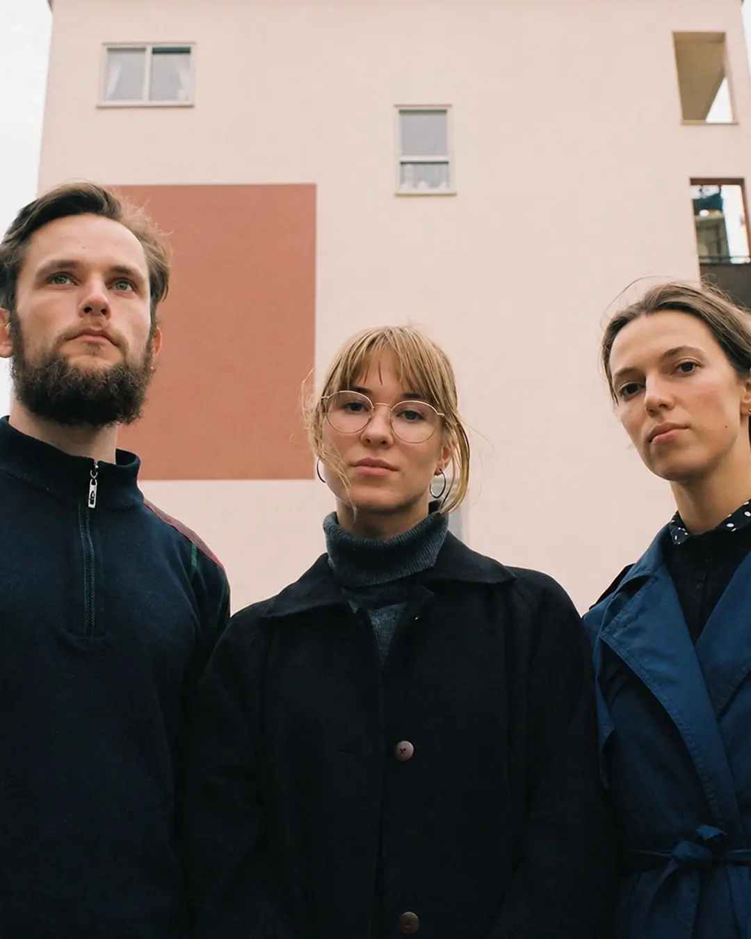 Drie bandleden van Meander poseren buiten voor een modern gebouw; twee vrouwen en een man kijken serieus in de camera.