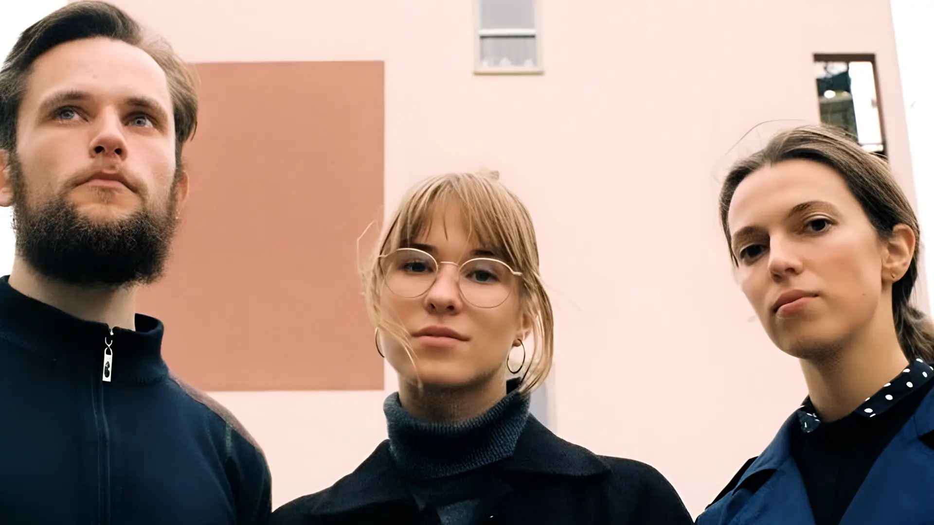 Drie bandleden van Meander poseren buiten voor een modern gebouw; twee vrouwen en een man kijken serieus in de camera.