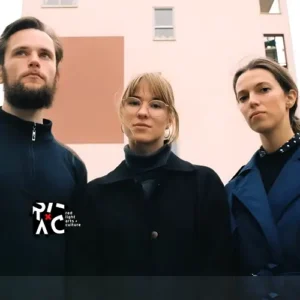 Drie bandleden van Meander poseren buiten voor een modern gebouw; twee vrouwen en een man kijken serieus in de camera.