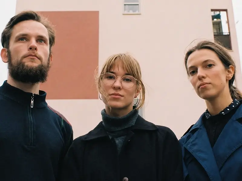 Drie bandleden van Meander poseren buiten voor een modern gebouw; twee vrouwen en een man kijken serieus in de camera.