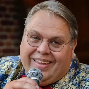 Portret van zanger Rico Parlevliet die glimlachend in een microfoon zingt, gekleed in een opvallend patroonjasje met strik, in een warme en intieme setting.