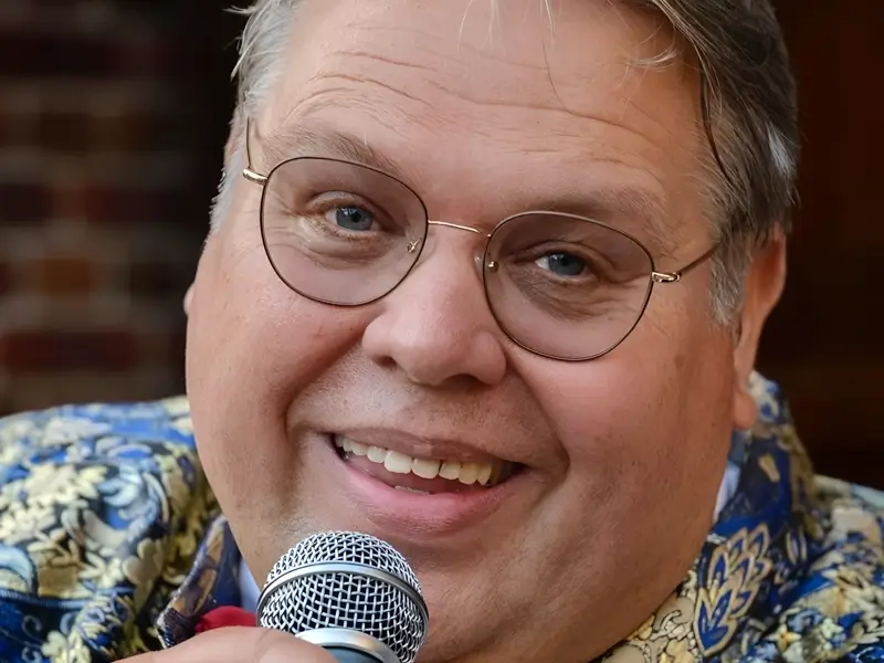 Portret van zanger Rico Parlevliet die glimlachend in een microfoon zingt, gekleed in een opvallend patroonjasje met strik, in een warme en intieme setting.
