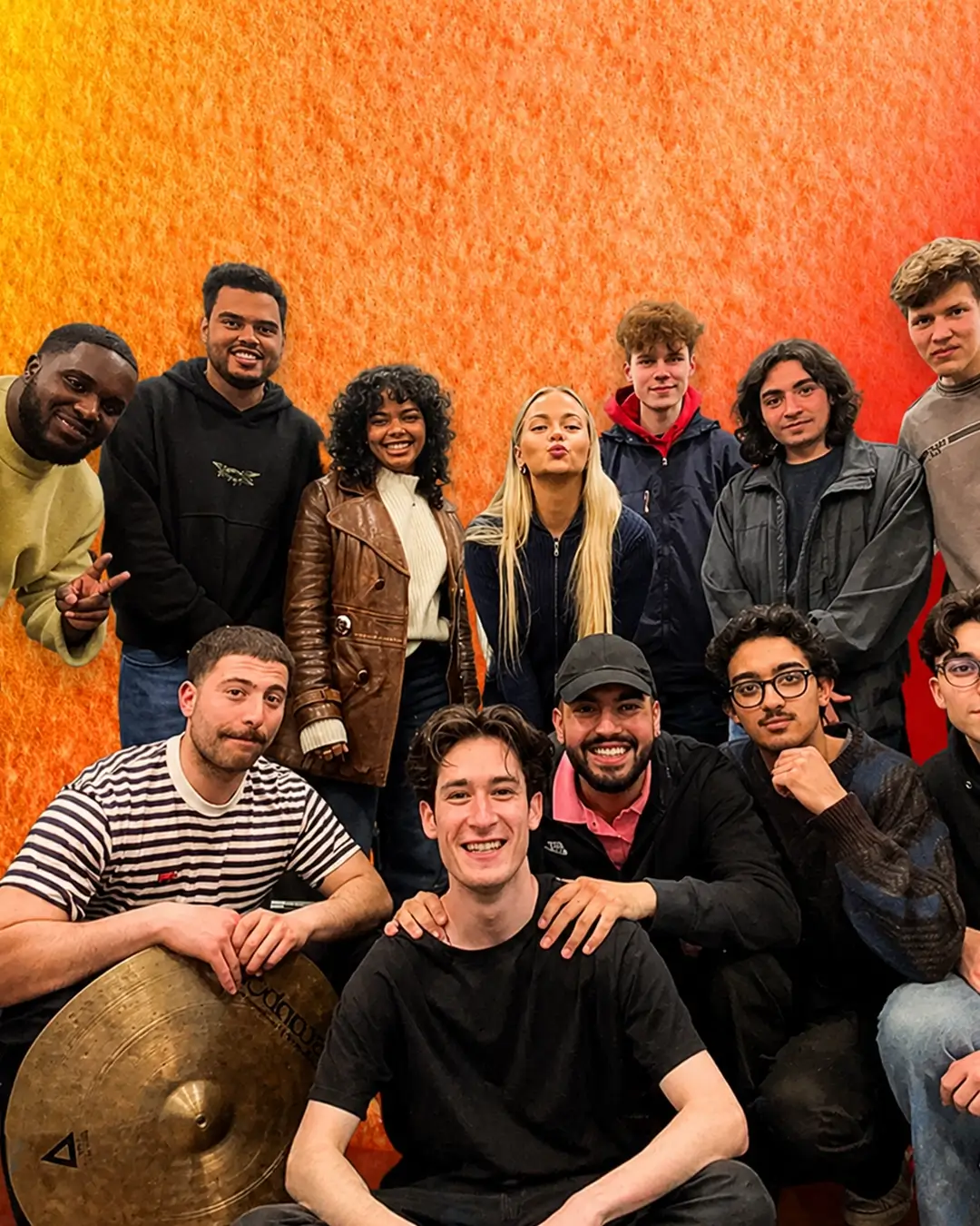 Een grote groep jonge muzikanten poseert samen voor een warme rood-oranje achtergrond, glimlachend en dicht bij elkaar, als bandfoto.