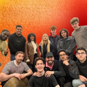 Een grote groep jonge muzikanten poseert samen voor een warme rood-oranje achtergrond, glimlachend en dicht bij elkaar, als bandfoto.