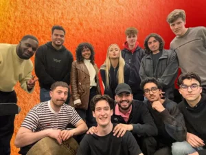 Een grote groep jonge muzikanten poseert samen voor een warme rood-oranje achtergrond, glimlachend en dicht bij elkaar, als bandfoto.