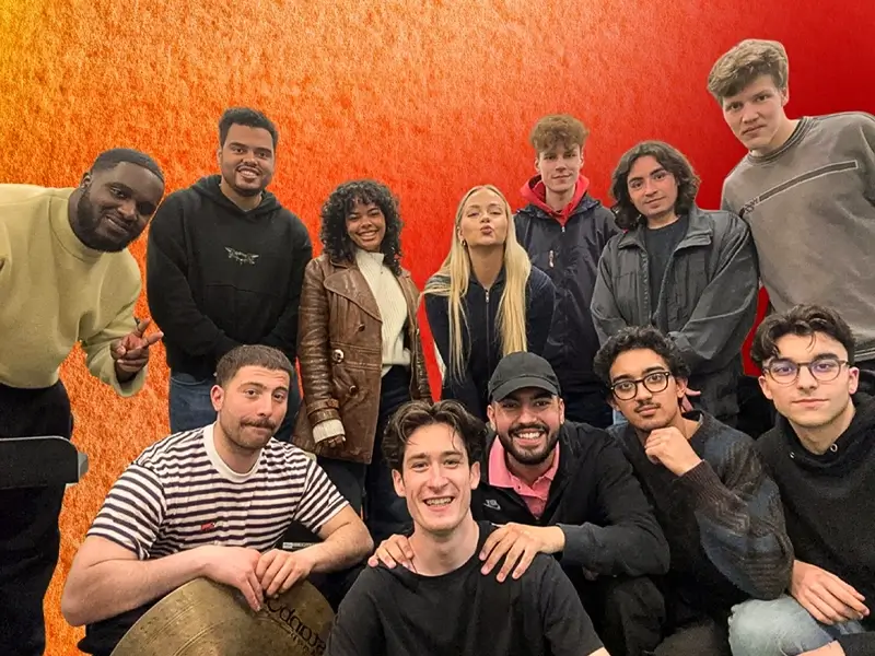 Een grote groep jonge muzikanten poseert samen voor een warme rood-oranje achtergrond, glimlachend en dicht bij elkaar, als bandfoto.