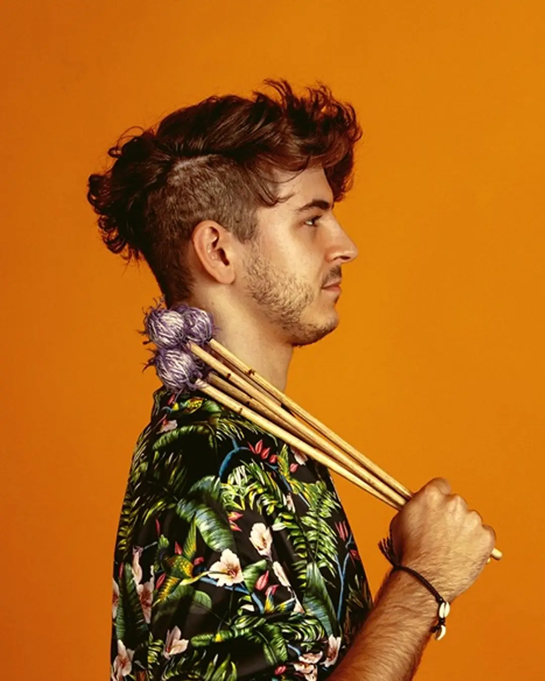 Een jonge man in profiel tegen een oranje achtergrond houdt vibrafoonstokken over zijn schouder, gekleed in een kleurrijk shirt.