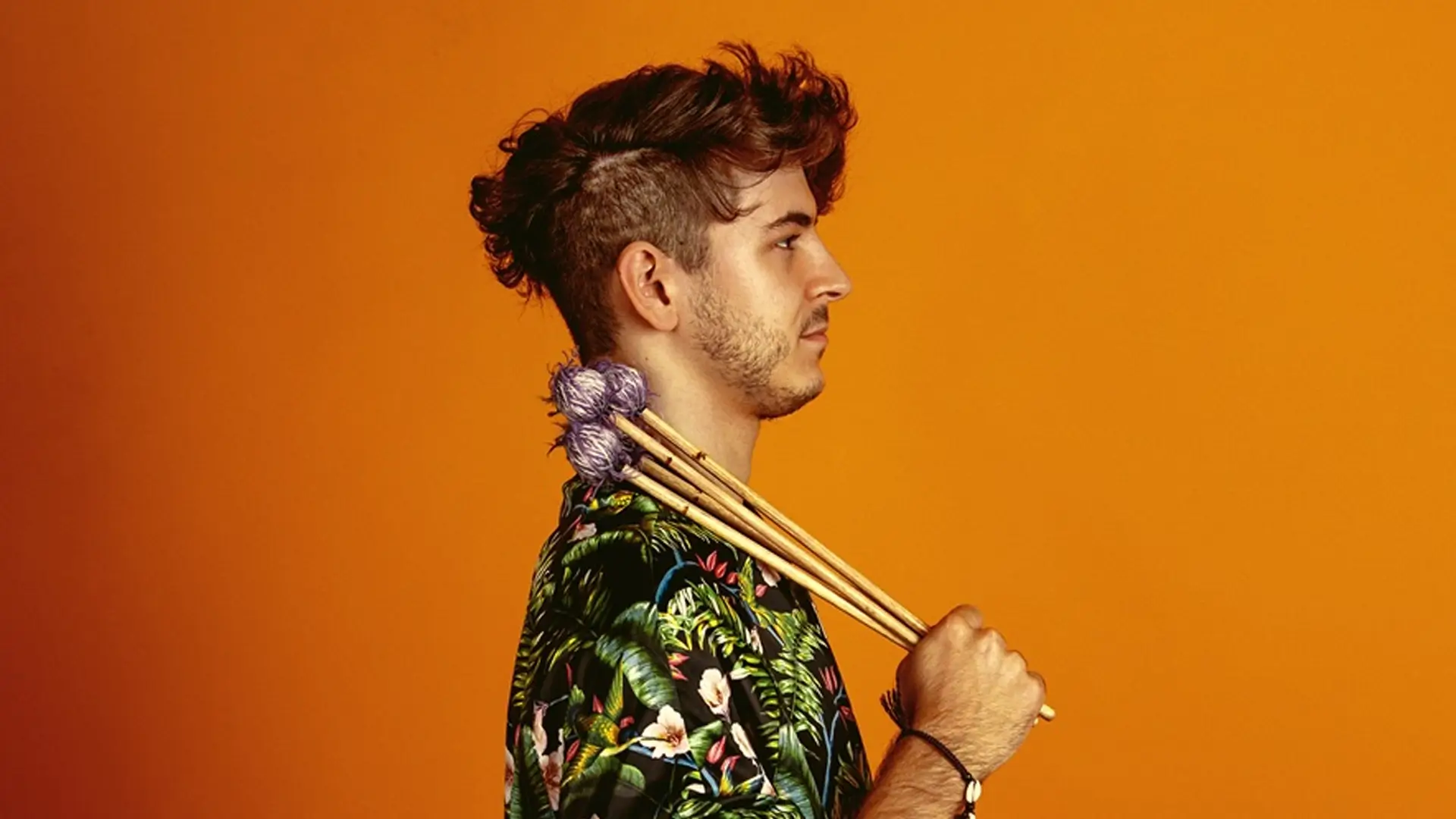 Een jonge man in profiel tegen een oranje achtergrond houdt vibrafoonstokken over zijn schouder, gekleed in een kleurrijk shirt.