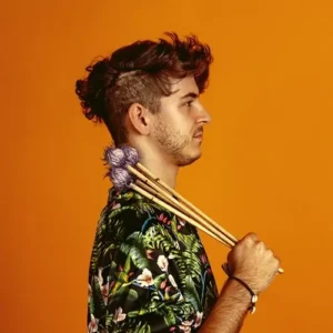 Een jonge man in profiel tegen een oranje achtergrond houdt vibrafoonstokken over zijn schouder, gekleed in een kleurrijk shirt.