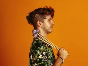Een jonge man in profiel tegen een oranje achtergrond houdt vibrafoonstokken over zijn schouder, gekleed in een kleurrijk shirt.