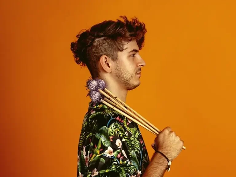 Een jonge man in profiel tegen een oranje achtergrond houdt vibrafoonstokken over zijn schouder, gekleed in een kleurrijk shirt.