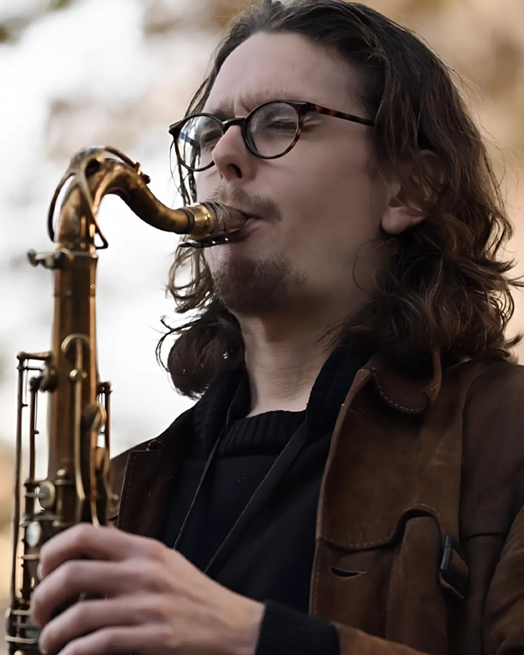 Een saxofonist speelt buiten op een tenorsaxofoon, met lang haar en een bril, gefocust op zijn spel tegen een zachte, onscherpe achtergrond.