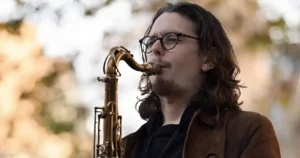 Een saxofonist speelt buiten op een tenorsaxofoon, met lang haar en een bril, gefocust op zijn spel tegen een zachte, onscherpe achtergrond.