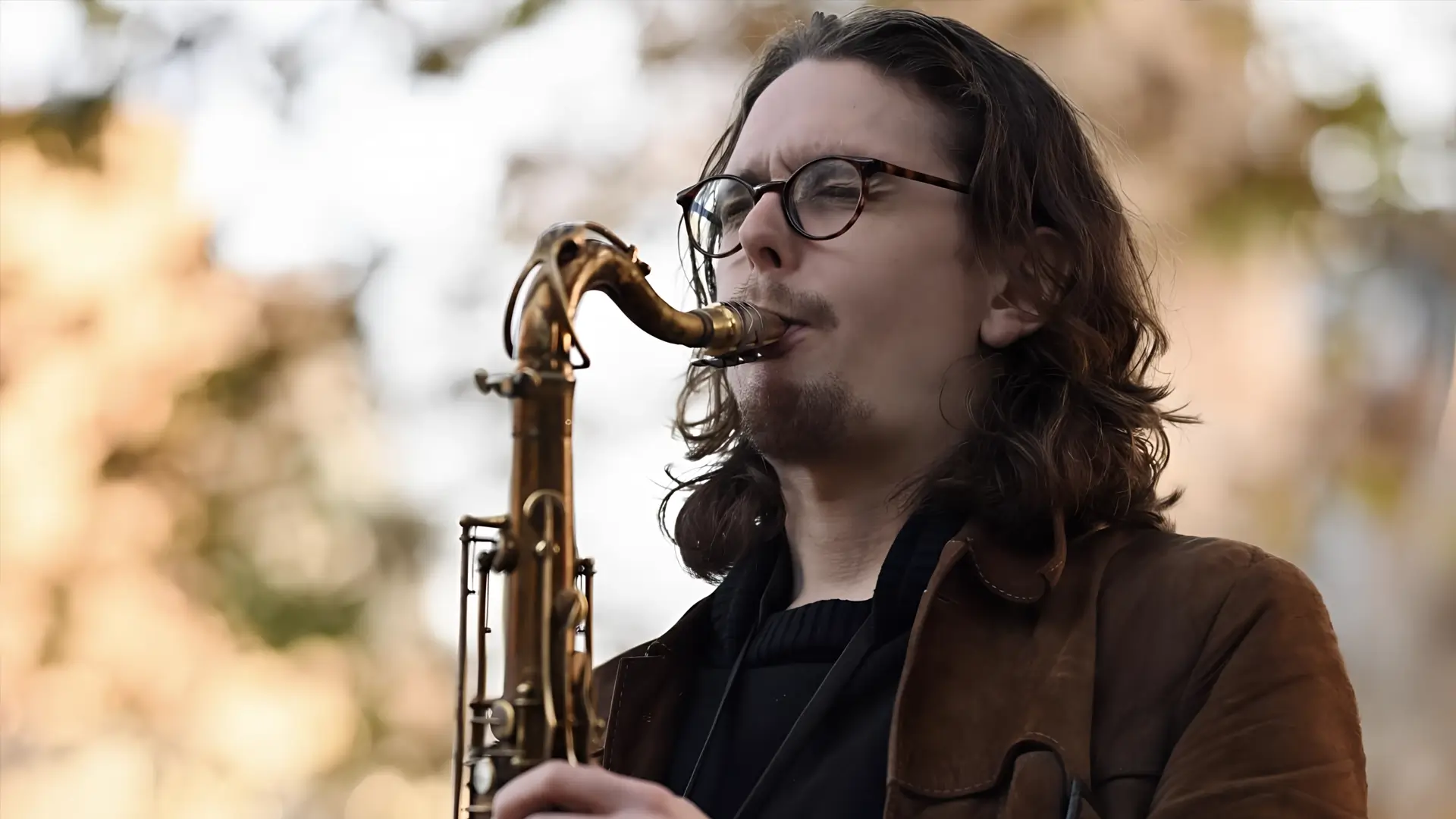Een saxofonist speelt buiten op een tenorsaxofoon, met lang haar en een bril, gefocust op zijn spel tegen een zachte, onscherpe achtergrond.