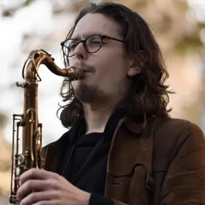 Een saxofonist speelt buiten op een tenorsaxofoon, met lang haar en een bril, gefocust op zijn spel tegen een zachte, onscherpe achtergrond.