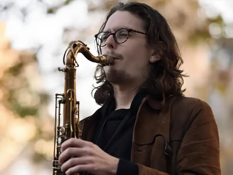 Een saxofonist speelt buiten op een tenorsaxofoon, met lang haar en een bril, gefocust op zijn spel tegen een zachte, onscherpe achtergrond.