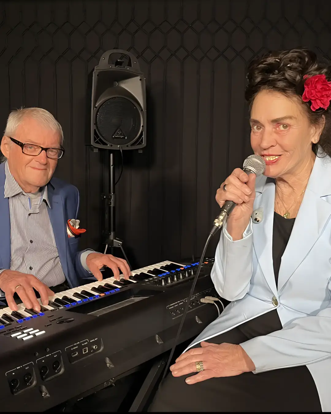 Een zangeres met een rode bloem in haar haar zingt in een microfoon terwijl een pianist naast haar keyboard speelt in een intieme theatersetting.