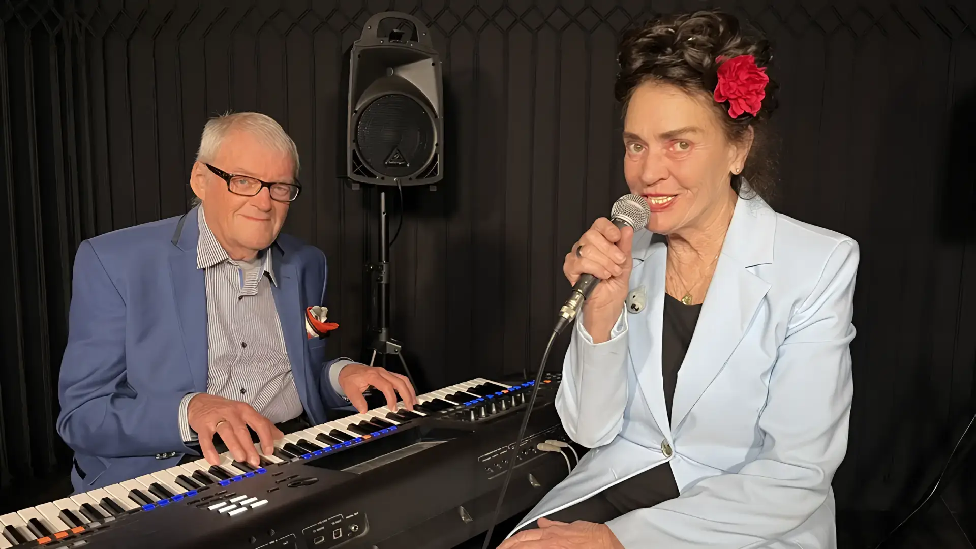 Een zangeres met een rode bloem in haar haar zingt in een microfoon terwijl een pianist naast haar keyboard speelt in een intieme theatersetting.