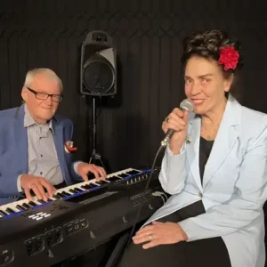 Een zangeres met een rode bloem in haar haar zingt in een microfoon terwijl een pianist naast haar keyboard speelt in een intieme theatersetting.