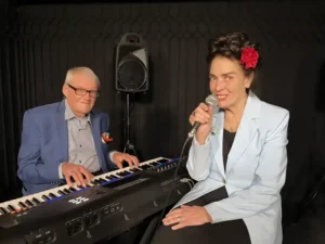 Een zangeres met een rode bloem in haar haar zingt in een microfoon terwijl een pianist naast haar keyboard speelt in een intieme theatersetting.