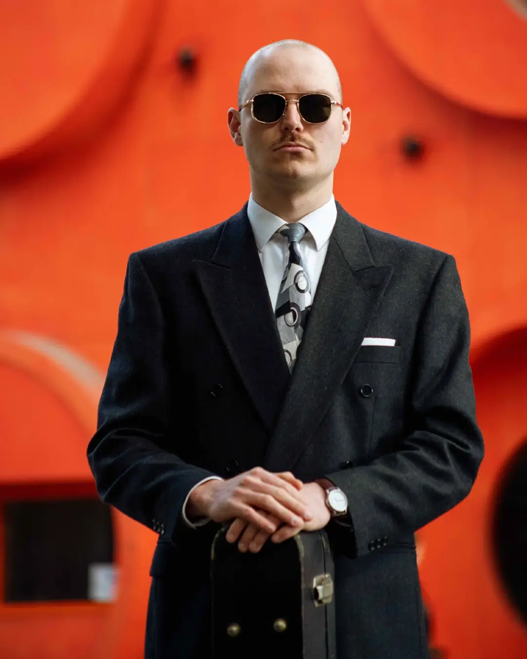 A sharply dressed man wearing sunglasses stands facing the camera with a case in his hands, set against a bright orange background with abstract shapes.