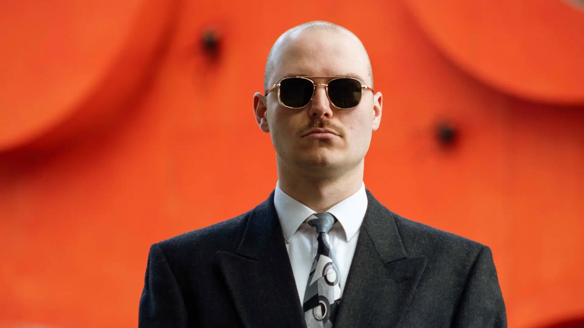 A sharply dressed man wearing sunglasses stands facing the camera with a case in his hands, set against a bright orange background with abstract shapes.