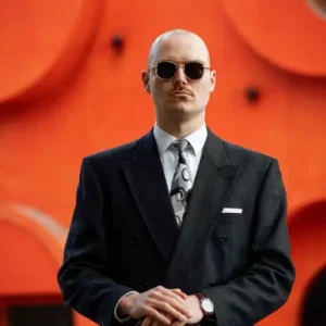 A sharply dressed man wearing sunglasses stands facing the camera with a case in his hands, set against a bright orange background with abstract shapes.