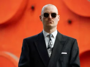 A sharply dressed man wearing sunglasses stands facing the camera with a case in his hands, set against a bright orange background with abstract shapes.