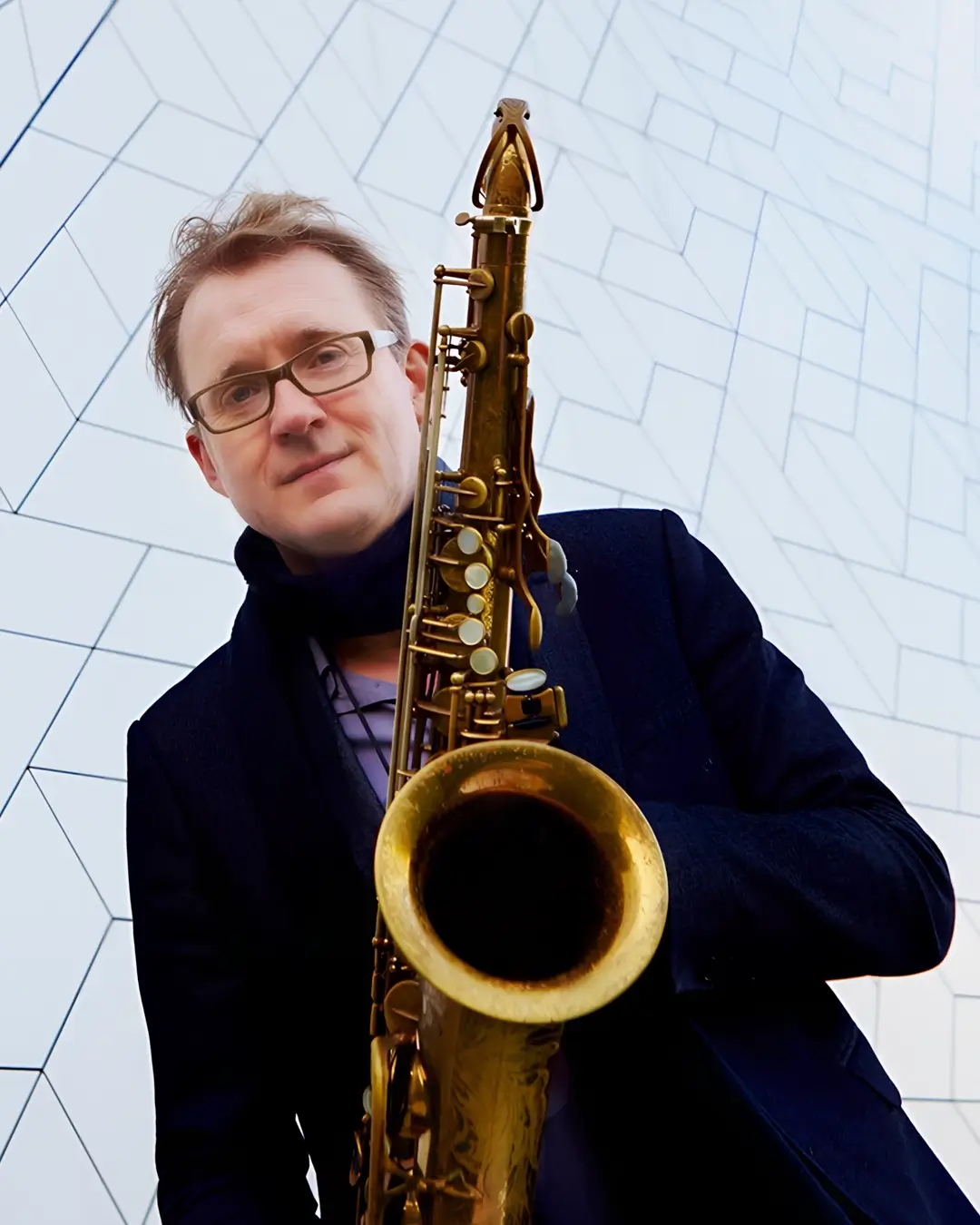 Portret van saxofonist Jasper Blom die een saxofoon vasthoudt en in de camera kijkt, gefotografeerd tegen een moderne geometrische gevel.