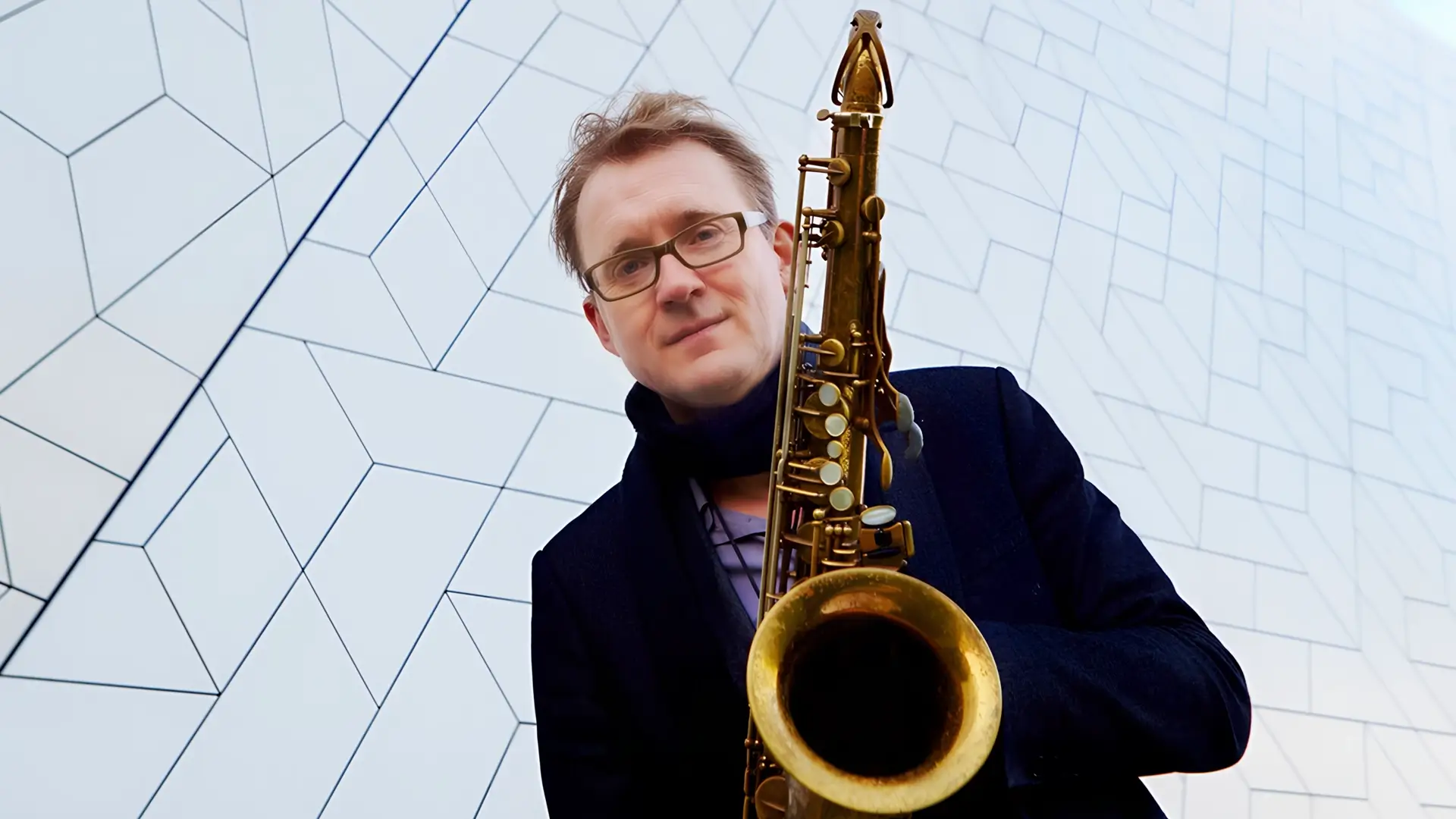 Portret van saxofonist Jasper Blom die een saxofoon vasthoudt en in de camera kijkt, gefotografeerd tegen een moderne geometrische gevel.