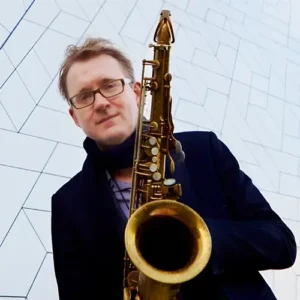 Portret van saxofonist Jasper Blom die een saxofoon vasthoudt en in de camera kijkt, gefotografeerd tegen een moderne geometrische gevel.