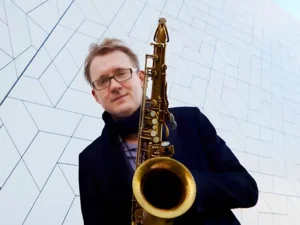 Portret van saxofonist Jasper Blom die een saxofoon vasthoudt en in de camera kijkt, gefotografeerd tegen een moderne geometrische gevel.