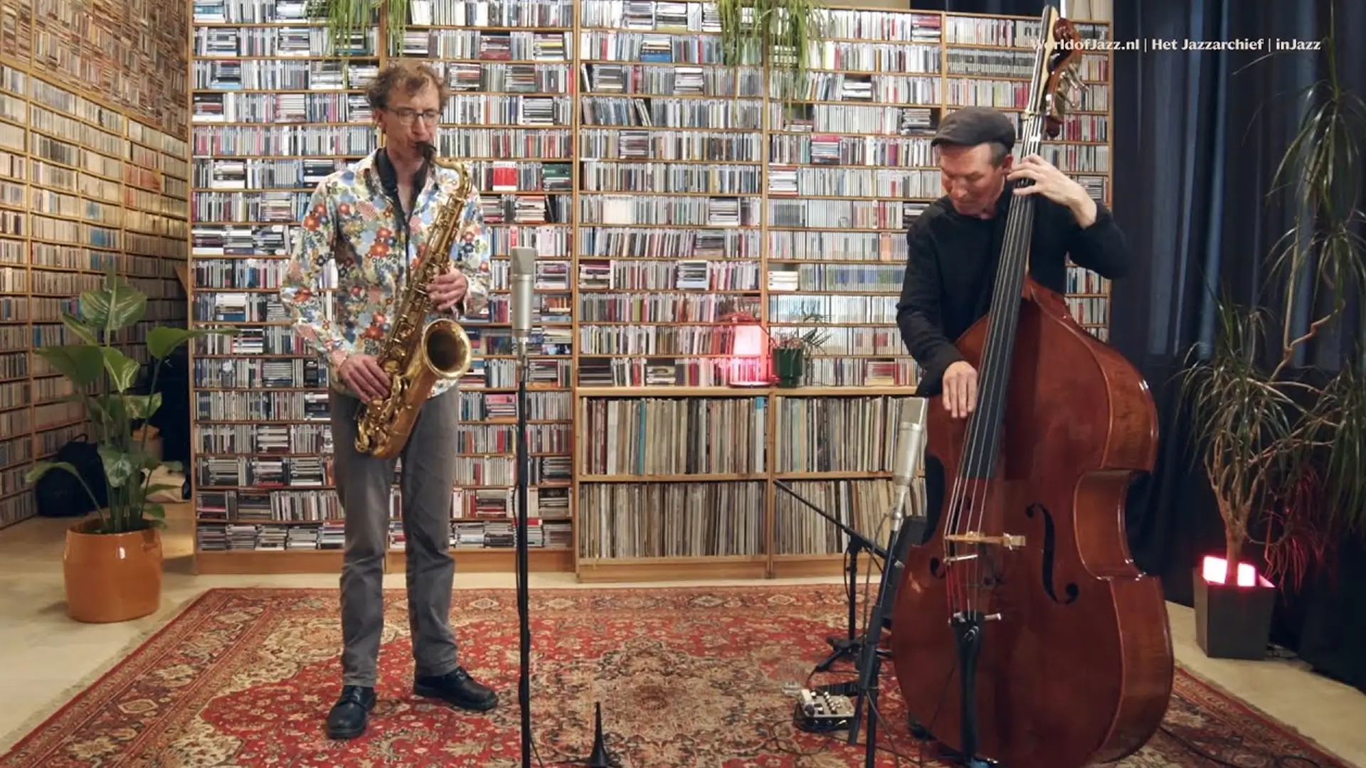 Een saxofonist en een contrabassist spelen een akoestisch jazzduo in een intieme ruimte, met een grote wand vol cd’s op de achtergrond en microfoons opgesteld voor hen.
