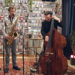 Een saxofonist en een contrabassist spelen een akoestisch jazzduo in een intieme ruimte, met een grote wand vol cd’s op de achtergrond en microfoons opgesteld voor hen.