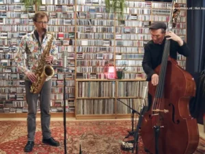 Een saxofonist en een contrabassist spelen een akoestisch jazzduo in een intieme ruimte, met een grote wand vol cd’s op de achtergrond en microfoons opgesteld voor hen.