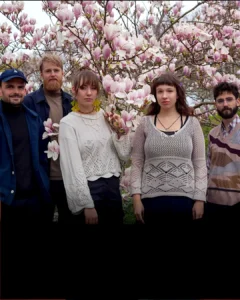 Five musicians stand together in front of a blooming magnolia tree, surrounded by pink flowers, looking calmly at the camera.